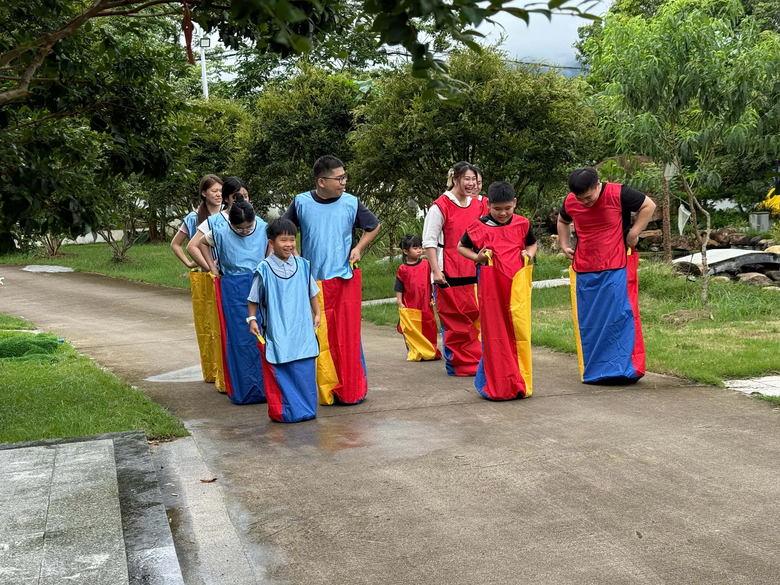 Adults and children in red and blue team bibs taking part in an outdoor sack race